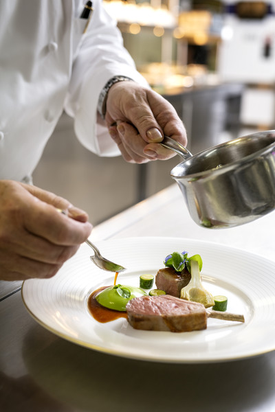 John Williams, Executive Chef at The Ritz London, elegantly plates a fine dining dish, adding a finishing touch of sauce with precision. The dish features beautifully presented meat, vibrant green purée, and delicate vegetables on a white plate.